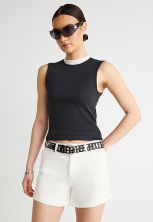 Kobieta w czarnym bezrękawniku, białych szortach z czarnym paskiem z ćwiekami, ciemnych okularach przeciwsłonecznych i srebrnej bransoletce, stojąca na jednolitym tle.