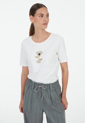 Frau mit braunen Haaren in einem niedrigen Pferdeschwanz, trägt ein weißes T-Shirt mit einem bestickten Martini-Glas und graue Nadelstreifenhose mit Kordelzug.