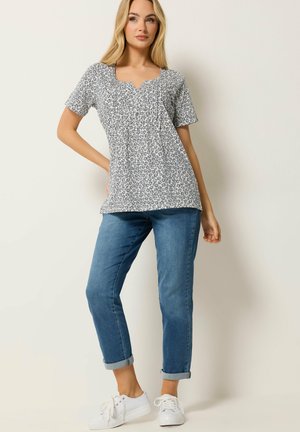 Femme debout, main sur la hanche, portant un haut fleuri noir et blanc à manches courtes, un jean bleu retroussé et des baskets blanches.