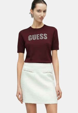 Donna che indossa una maglia color bordeaux a maniche corte con il testo "GUESS", abbinata a una minigonna bianca texturizzata con piccole tasche frontali.