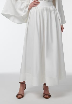 Persona con una falda larga blanca, blusa blanca de manga ancha y sandalias marrones de punta abierta, de pie frente a un fondo liso.