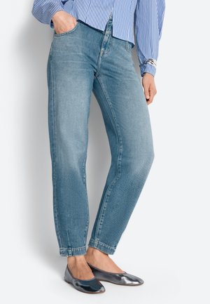 Person iført lyseblå jeans, metallisk sølvfarvede flade sko og en blå- og hvidstribet skjorte med hånden i lommen.