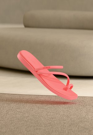 Sandaletto rosa corallo senza lacci con anello per il dito e sottili cinturini, sospeso sopra un tappeto beige testurizzato con sfondo sfocato di un divano beige.