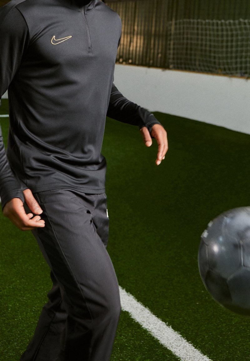 Schwarzes Nike Sportoberteil mit halber Reißverschluss-Kragen, kombiniert mit schwarzen Hosen. Glatte Textur, enganliegendes Design, mit goldfarbenem Logo-Akzent auf der Brust.