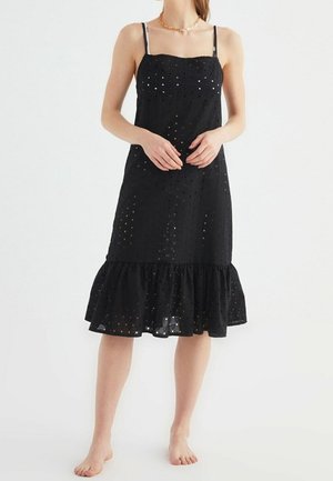 Kobieta w czarnej, bezrękawnikowej sukience midi z ażurowym wzorem i falbaną u dołu, stojąca boso na jednolitym tle.