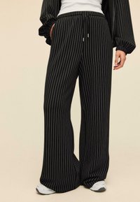 Mustat pinstripe-housut leveillä lahkeilla, joustavalla vyötäröllä, sivutaskuilla ja teksturoidulla kankaalla. Yhdistetty valkoisiin tennareihin.