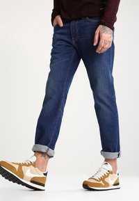 Vaqueros de mezclilla azul con corte ajustado, puños enrollados y detalles de costura sutiles, combinados con zapatillas Nike beige y blancas.