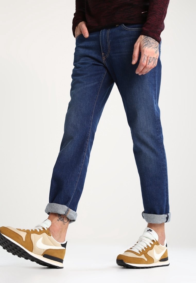 Vaqueros de mezclilla azul con corte ajustado, puños enrollados y detalles de costura sutiles, combinados con zapatillas Nike beige y blancas.