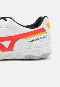 Zapatilla blanca con acentos rojos y amarillos, parte superior sintética texturizada, forro negro y suela de goma con un patrón para agarre.