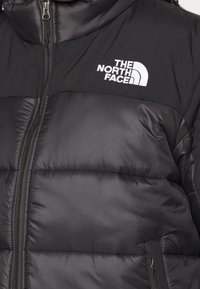 Veste matelassée noire avec un col haut, dotée d'une fermeture éclair à l'avant et du logo blanc "THE NORTH FACE" sur le côté supérieur gauche.
