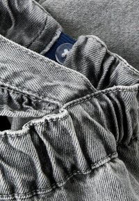 Tessuto in denim grigio con dettaglio a vita arricciata, caratterizzato da cuciture visibili e chiusura con bottone. La texture è leggermente consumata.