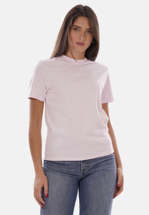 Donna con capelli castani e lunghi che indossa una t-shirt Calvin Klein rosa chiaro e jeans blu, in piedi davanti a uno sfondo bianco semplice.
