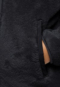 Schwarze Fleecejacke aus weichem, strukturiertem Stoff, mit einem kontrastierenden, glatten Besatz entlang der Taschen und Ärmelkanten.