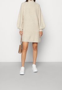 Robe en maille beige avec un col montant et des manches ballon, associée à des baskets blanches et un petit sac à main marron. Texture lisse, longueur genoux.