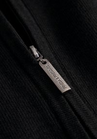 Zwarte ritsdetail op een gestructureerde stof, met een zilveren metalen trekker met reliëfbranding "FRIEDA & FREDDIES." Close-up afbeelding.