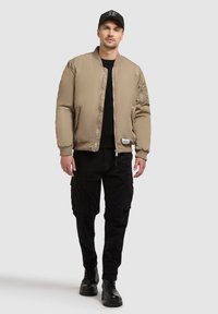 Béžová bomber bunda s rebrovaným golierom, zipsom a bočnými vreckami, nosená cez čiernu košeľu a čierne cargo nohavice, doplnená čiernymi topánkami.