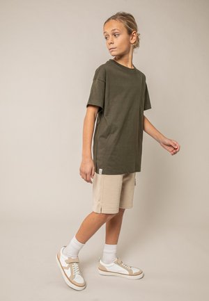 Jong persoon in olijfgroen T-shirt, beige korte broek, witte sokken en sneakers staand en zijwaarts kijkend tegen een effen achtergrond.