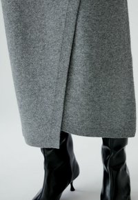 Jupe tricotée grise avec une fente latérale, dotée d'un ourlet côtelé. Associée à des bottes noires jusqu'aux genoux, mettant en valeur la texture du cuir lisse.