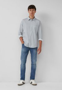 Junger Mann steht an einer weißen Wand und trägt ein hellgraues Hemd mit Knöpfen, eine blaue Jeans und weiße Sneakers mit schwarzen Streifen.