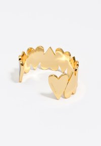 Brazalete de pulsera en forma de corazón de tono dorado con un acabado suave y brillante. Presenta formas alternas de corazón y zigzag a lo largo de la banda.