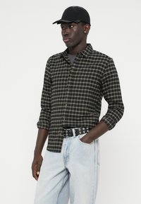 Μαύρο και ελιά κρουαζέ πουκάμισο με κουμπιά μπροστά, γυρισμένα μανίκια, φορεμένο με ανοιχτό μπλε τζιν και μια μαύρη ζώνη με ατσάλινες καρφίτσες. Φοράει μαύρο καπέλο.