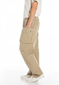Pantalones cargo beige con cintura elástica, bolsillos laterales y un bolsillo con solapa en el muslo; hechos de una tela ligera con un acabado texturizado.