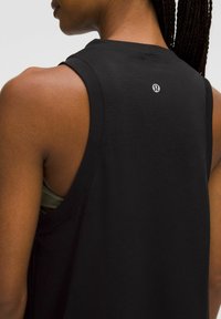 Close-up van een persoon met gevlochten haar die een zwart mouwloos sporttop draagt met een klein reflecterend logo op de bovenrug.