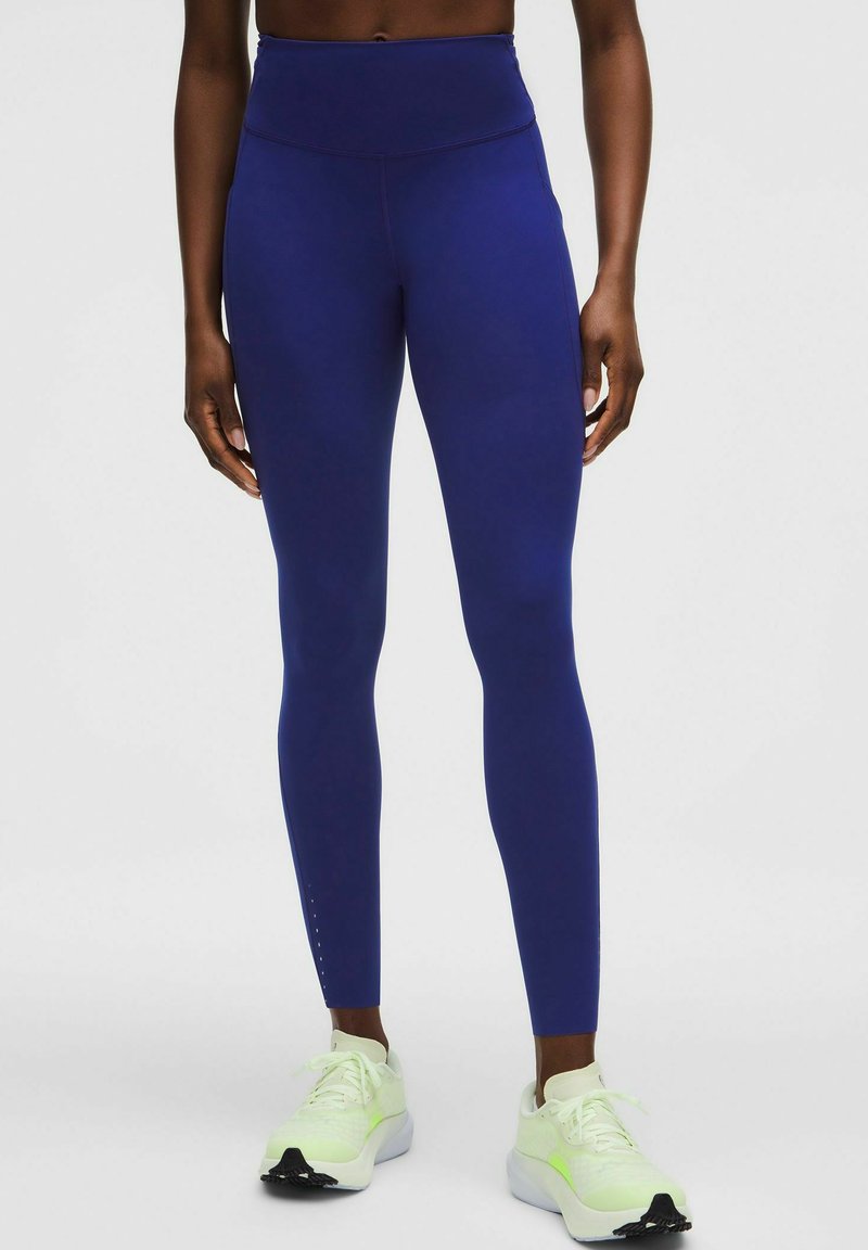 Person iført højtaljede, mørkeblå leggings og off-white løbesko med neongrønne accenter, stående mod en ensfarvet lys baggrund.
