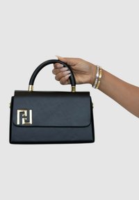 Bolso de cuero negro con herrajes en tono dorado, forma estructurada, asa superior y un acento de logo en la parte delantera. Textura suave.