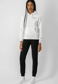 Sudadera de felpa blanca con un bolsillo frontal y cordón ajustable, combinada con pantalones jogger negros y zapatillas blancas.