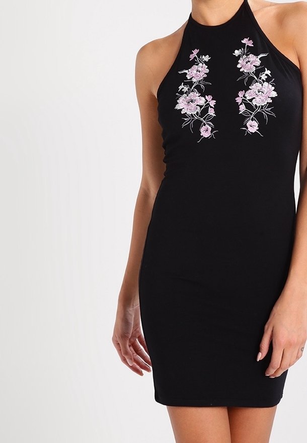 Robe noire sans manches à col licou avec broderie florale rose sur le haut devant, portée par une personne les mains détendues le long du corps.