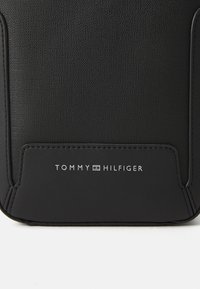 Crna teksturirana platnena torba s glatkom crnom površinom koja na dnu sadrži bijelu oznaku "TOMMY HILFIGER". Minimalistički dizajn.