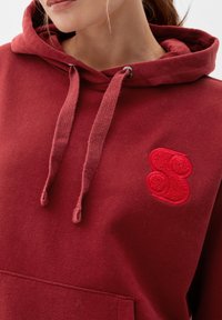 Sweat à capuche bordeaux en coton doux, avec une poche avant, une capuche à cordon de serrage et un patch logo rouge texturé sur la poitrine gauche.