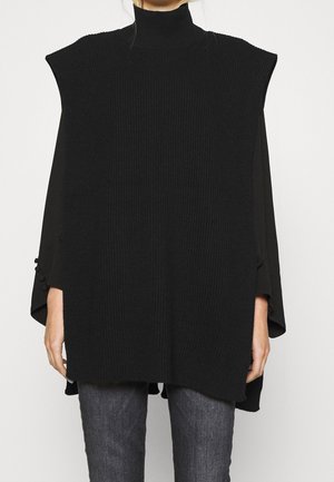 Vrouw die een zwarte geribbelde gebreide mouwloze coltrui draagt over een zwarte longsleeve en donkergrijze spijkerbroek, staand tegen een witte achtergrond.