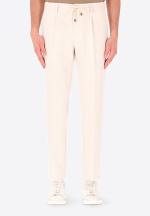 Uomo che indossa pantaloni crema a righe sottili con vita con coulisse e scarpe da ginnastica bianche, in piedi con le mani rilassate lungo i fianchi.