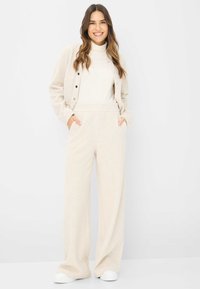 Beige Strickjacke mit Knopfverschluss, kombiniert mit hoch taillierten, weit geschnittenen beigen Hosen. Weicher Stoff und zwei Seitentaschen. Weiße Schuhe.