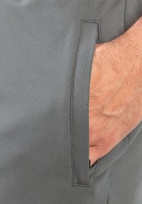 Grijze, rekbare stof van broek met een zijzak. Textuur lijkt glad, enkele gestikte naden zichtbaar. Hand gedeeltelijk in de zak gestoken.