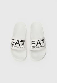 Bele natika z belo površino, ki imajo širok trak. Črn logotip "EA7 EMPORIO ARMANI" je natisnjen čez trak. Preprosta zasnova.