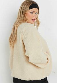 Kremowa polarowa kurtka o miękkiej teksturze, luźnym kroju, opadających ramionach i bocznych kieszeniach. Lekko zaokrąglony dół i długie rękawy.