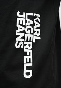 Μαύρο ύφασμα με έντονα λευκά κάθετα γράμματα που γράφουν «KARL LAGERFELD JEANS» και μια μικρή σιλουέτα ενός ατόμου που σκαρφαλώνει στο γράμμα «R».