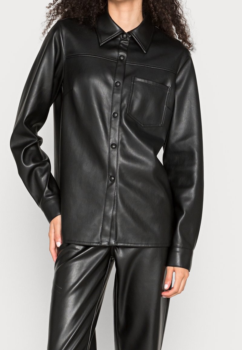 Femme portant une chemise noire en simili cuir boutonnée avec une poche poitrine et un pantalon assorti en simili cuir, main posée sur la hanche.