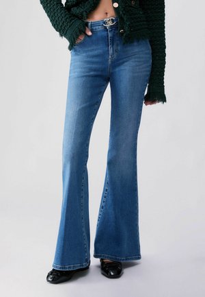Personne portant un jean bleu évasé taille mi-haute, des chaussures noires et une veste verte texturée à manches à franges, debout devant un fond uni.