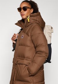 Chaleco acolchado marrón con capucha, con un cordón amarillo, bolsillos laterales y un emblema. Se puede ver una mochila de color claro y con un patrón.