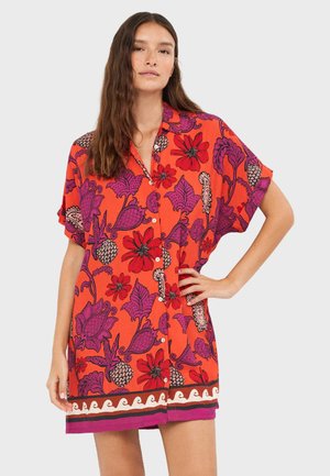 Frau trägt ein locker sitzendes Knopfleistenkleid mit kurzen Ärmeln und rotem, lila Blumendesign sowie Ananasmuster, steht mit einer Hand in der Hüfte.