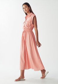 Rožinė be rankovių suknelė su apykakle, elastinga juosmens juosta, dviem krūtinės kišenėm ir laisvai krentančia A formos sijonu. Derinama su minimalistiniais sandalais.