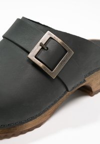 Sort læderklump med metalspænde, træsål og syet detalje langs kanten. Glat tekstur og rund tå-design.