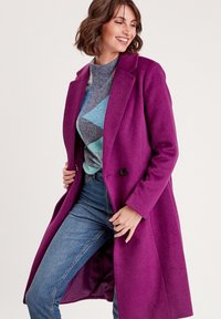 Manteau en laine magenta au design ajusté, avec deux boutons et un col cranté, associé à un pull à motifs et un jean bleu.