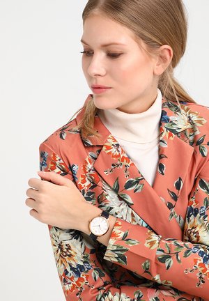 Femme aux cheveux blonds attachés en queue de cheval, portant un col roulé blanc et un blazer orange à motif floral, regardant sa montre-bracelet à bracelet noir.