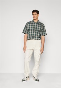 Chemise à manches courtes à carreaux vert et beige, dotée de deux poches poitrine. Portée avec un pantalon couleur crème et des baskets à motifs.