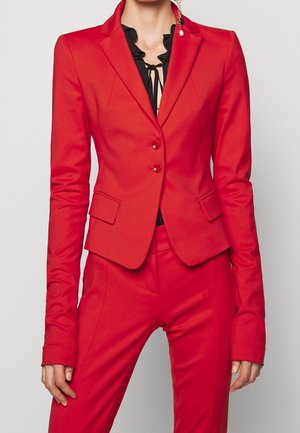 Vrouw draagt een nauwsluitend rood colbert en bijpassende broek met een zwart gerimpeld blouse eronder, zichtbaar van nek tot midden bovenbeen.